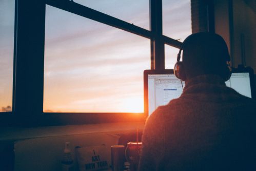 Homem trabalhando em uma mesa de computador com fone de ouvido virado para um janela onde se vê o por do sol